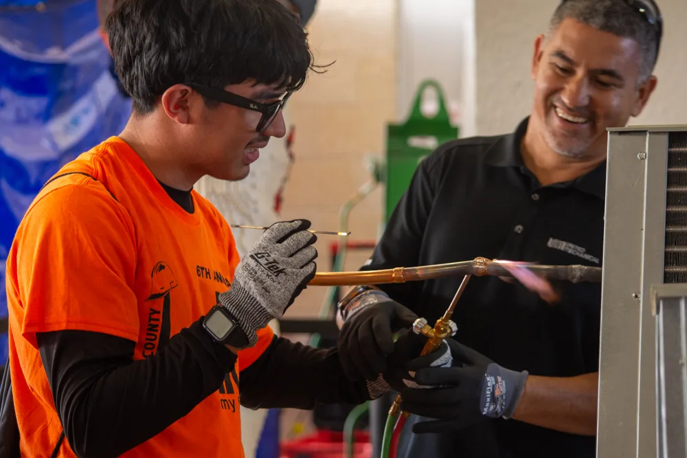 A student in an orange Build My Future shirt and work gloves holds a torch to copper pipe beside a smiling instructor next to an air conditioning unit.