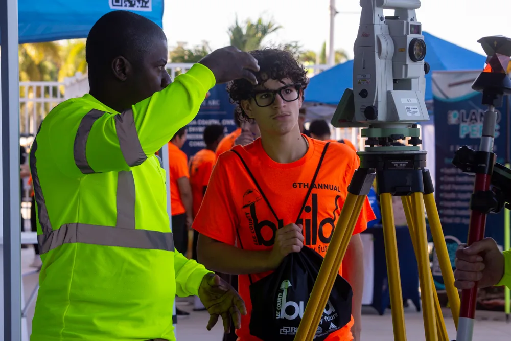 A worker in a high-visibility shirt shows a student in an orange Build My Future shirt how to use a surveying instrument on a yellow tripod.