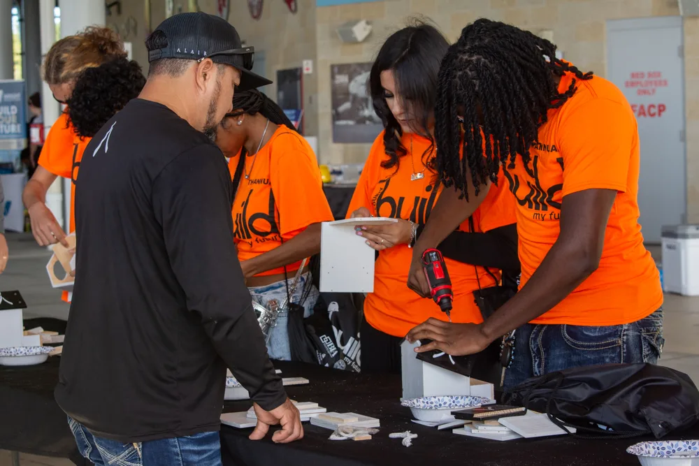 Students in orange Build My Future shirts use a cordless drill to assemble small wooden projects at a table.