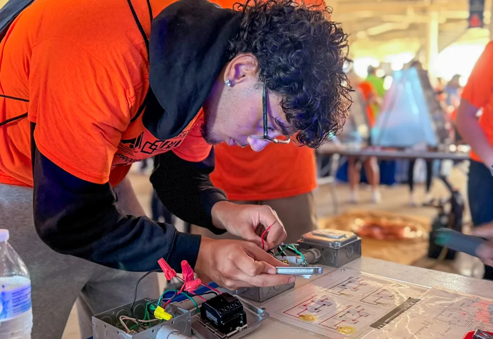 A student in an orange shirt connects colored wires inside an electrical box at a table with wiring diagrams.