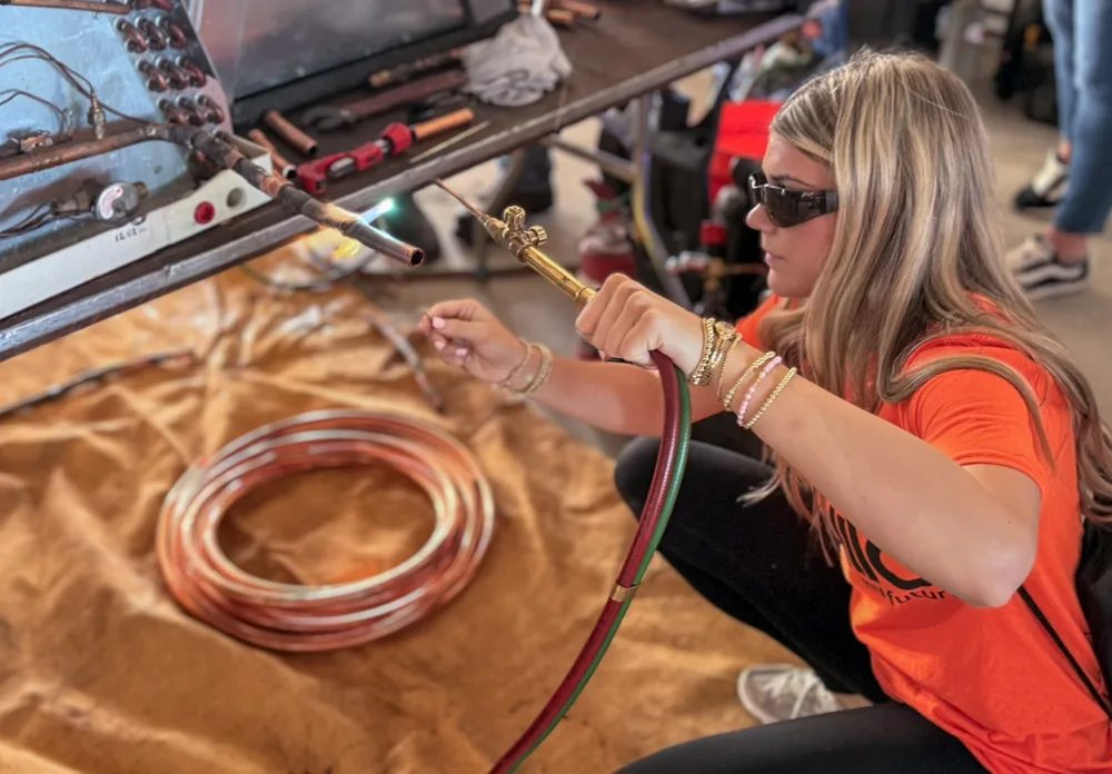 A student in sunglasses and an orange shirt kneels to hold a torch to copper fittings on a training rig, with a coil of copper tubing on the floor.