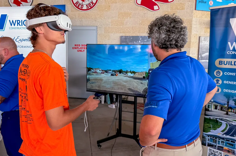 A student wearing a virtual reality headset stands in front of a screen showing a construction site, beside a man in a blue shirt at the Wright Construction Group stand.