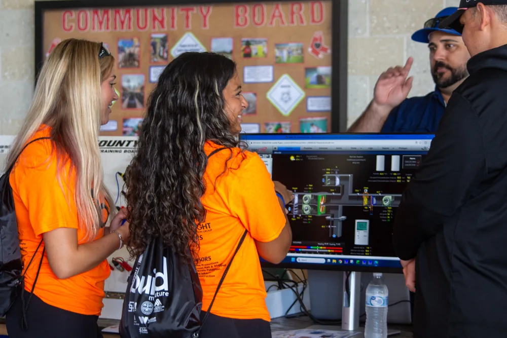 Two students in orange Build My Future shirts look at a computer screen showing a building systems control display while two men explain it.