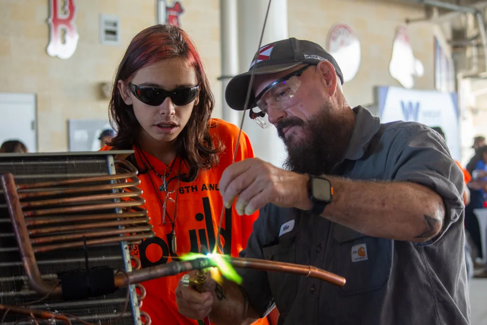A student in a Build My Future shirt watches a tradesman work a torch on copper tubing at JetBlue Park.